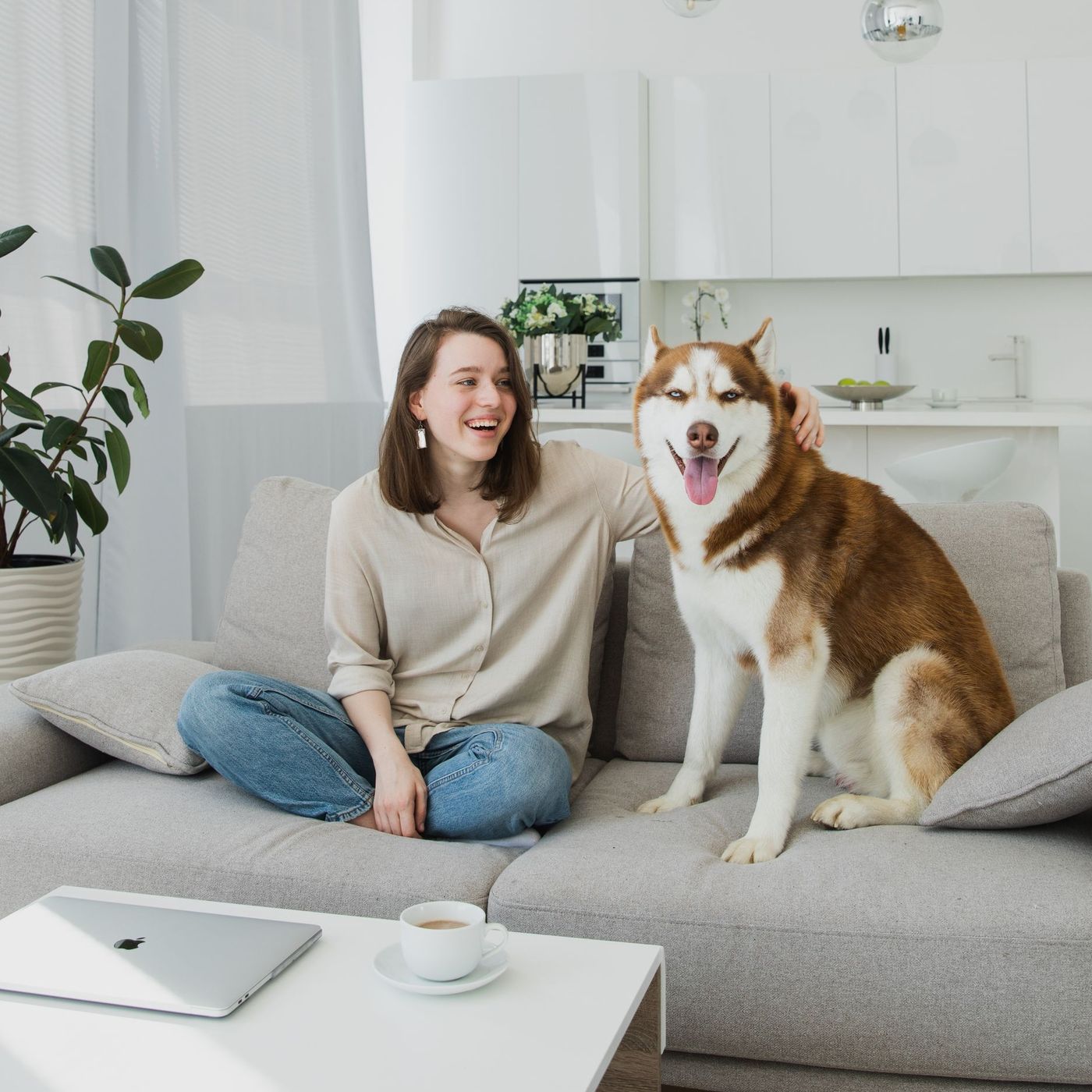 A smiling woman sits on the sofa next to a large husky in a bright living room.